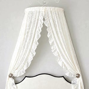 May include: A white bed canopy featuring sheer, ruffled fabric panels that drape downward from a circular mount. The canopy is accented with a crystal-like beaded trim at the top and two circular silver-toned tie-backs holding the curtains.