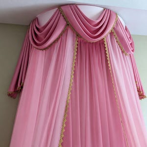 May include: A pink bed canopy featuring a semi-circular top mount with draped fabric swags. The canopy includes sheer pink curtains and opaque pink side panels, all accented with decorative gold lace trim along the edges and the valance.