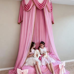 May include: A pink bed canopy featuring a circular top mount with draped fabric panels, gold trim accents, and two large decorative bows attached to the lower corners of the bed skirt. The sheer pink curtains hang from the ceiling to the floor.