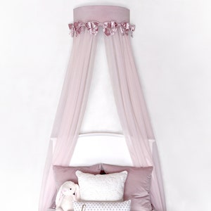 May include: A pink canopy bed with a white headboard and a pink velvet trim. The canopy is made of sheer fabric and hangs from a circular frame.
