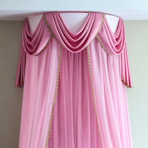 May include: A pink bed canopy featuring a semi-circular top frame with draped fabric swags. The canopy includes sheer pink curtains hanging vertically, accented with gold-coloured decorative trim along the edges and the draped sections.