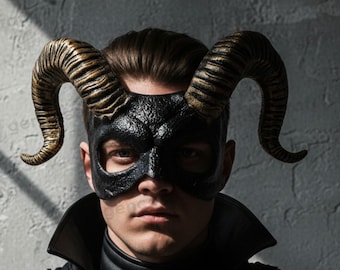 May include: A black mask featuring large, textured, curved horns with gold-toned highlights. The mask covers the upper face and is worn with a black layered garment featuring shoulder plates, multiple straps, and metallic studs across the chest.