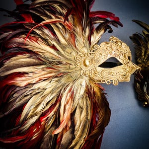 Puede incluir: Una m&aacute;scara veneciana dorada con intrincados detalles de encaje y una joya central. La m&aacute;scara est&aacute; adornada con un abanico de plumas en tonos rojos, dorados y marrones. Una segunda m&aacute;scara es visible en el fondo.