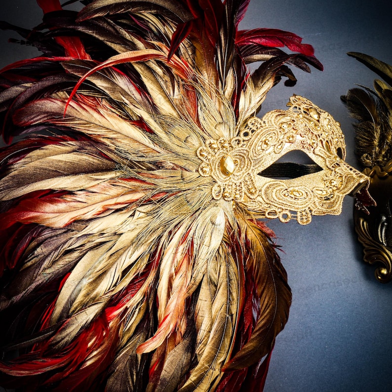 Puede incluir: Una m&aacute;scara veneciana dorada con intrincados detalles de encaje y una joya central. La m&aacute;scara est&aacute; adornada con un abanico de plumas en tonos rojos, dorados y marrones. Una segunda m&aacute;scara es visible en el fondo.