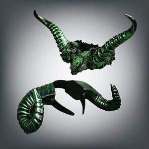 May include: Two green, ornate horns with a dark base. The top horn features floral detailing and a textured surface. The lower horn has a spiral design on one end and a curved shape. The horns are set against a gradient gray background.