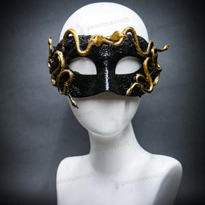 Puede incluir: Una máscara negra de carnaval con serpientes doradas enrolladas en los bordes. La máscara tiene una superficie texturizada y se muestra en una cabeza de maniquí blanca. Las serpientes están intrincadamente detalladas.
