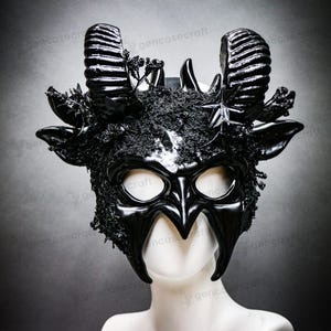Könnte beinhalten: Eine schwarze Maske mit Hörnern und glänzendem Finish, mit großen, gebogenen Hörnern und komplizierten Details. Die Maske hat Augenöffnungen und ist mit strukturierten, dunklen Verzierungen versehen, die eine dramatische und gotische Ästhetik erzeugen.