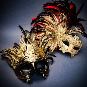 Puede incluir: Dos m&aacute;scaras de carnaval ornamentadas. Una es dorada con plumas negras y un dise&ntilde;o decorativo. La otra es dorada con plumas rojas y negras y un dise&ntilde;o de encaje. Ambas m&aacute;scaras tienen aberturas para los ojos.