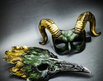 May include: Two handcrafted masks. One is a horned mask with gold-colored horns and a green and black base. The other is a bird-like mask with green and gold feathers and a long beak. Both masks are on a gray surface.