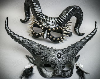 May include: Two black masquerade masks with horns. One mask has a black lace design and a single black jewel. The other mask has a spiked design with gold accents.