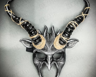 May include: A silver and black horned mask with a textured finish. The mask features large, curved horns wrapped with twine. The mask has eye and mouth openings and an elastic strap for wear.