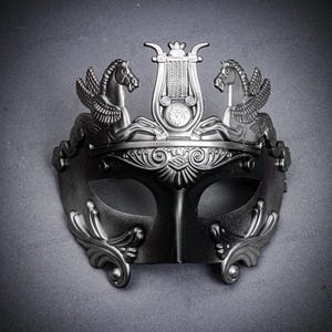 May include: A silver-colored masquerade mask with a decorative design. The mask features two winged horses flanking a lyre, with ornate scrollwork and eye openings. The mask is likely made of metal or a similar material.