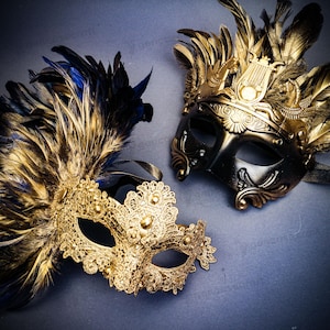 May include: Two ornate masquerade masks. One is gold with blue and gold feathers, the other is black and gold. Both masks have intricate designs and are suitable for a masquerade ball or costume party.