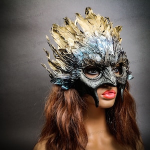 Könnte beinhalten: Eine schwarze Maskenballmaske mit goldenen und blauen Federn. Die Maske hat ein vogelartiges Design mit einem spitzen Schnabel und großen Augen.
