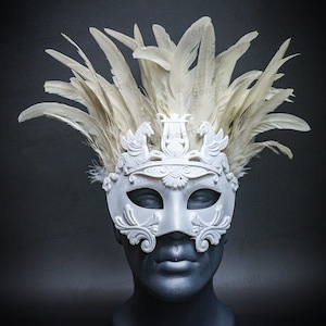May include: A white masquerade mask with a decorative design, featuring a crown with two winged horses and a lyre, and adorned with cream-colored feathers. The mask is displayed on a dark mannequin head.