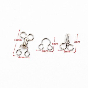 May include: Three silver metal hook and eye closures. The largest closure measures 11 mm long and 6 mm wide. The smaller closures measure 7 mm long and 6 mm wide. The smallest closure measures 5 mm long and 6 mm wide.