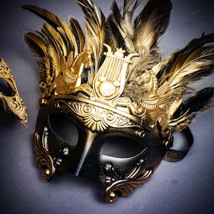 Puede incluir: Una m&aacute;scara de carnaval negra y dorada con detalles ornamentados y plumas doradas. La m&aacute;scara presenta una corona dorada con dise&ntilde;os de caballos alados y lira. Una segunda m&aacute;scara es parcialmente visible en el fondo.