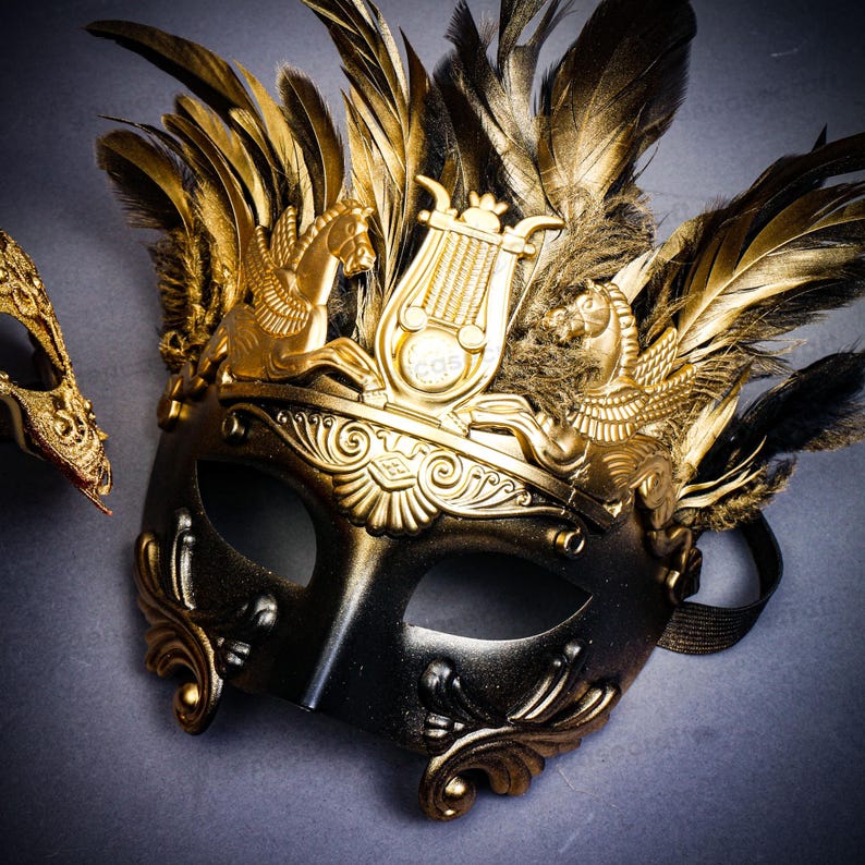 Puede incluir: Una m&aacute;scara de carnaval negra y dorada con detalles ornamentados y plumas doradas. La m&aacute;scara presenta una corona dorada con dise&ntilde;os de caballos alados y lira. Una segunda m&aacute;scara es parcialmente visible en el fondo.