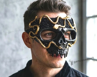 May include: A black skull mask featuring gold-colored snake motifs that wrap around the eye sockets and forehead. The mask has a textured black base with jagged, tooth-like details along the bottom edge, covering the upper face area.