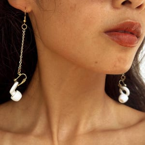 Op de afbeelding: Oorbellen met witte, oortelefoonvormige hangers die aan goudkleurige haken en schakels hangen. De oorbellen worden gedragen, met één zichtbaar aan elk oor. Achtergrond: gezicht en nek.