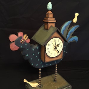 May include: A wooden rooster clock with a blue and green painted design. The clock face is white with black numbers and hands. The rooster is standing on a base with the text "Always time to help those in need".