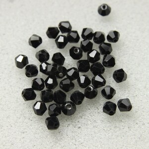 Puede incluir: Un grupo de cuentas de vidrio facetadas negras, esparcidas sobre una superficie blanca. Las cuentas son pequeñas y tienen un acabado brillante.