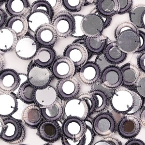 Peut inclure: Gros plan sur un tas de rondelles argentées et grises ressemblant à des miroirs avec une bordure en crochet gris foncé. Les rondelles sont éparpillées sur une surface blanche.
