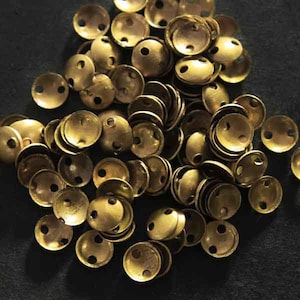 Puede incluir: Un montón de cuentas de metal de color dorado con dos agujeros cada una. Las cuentas son redondas y tienen una forma ligeramente abovedada.