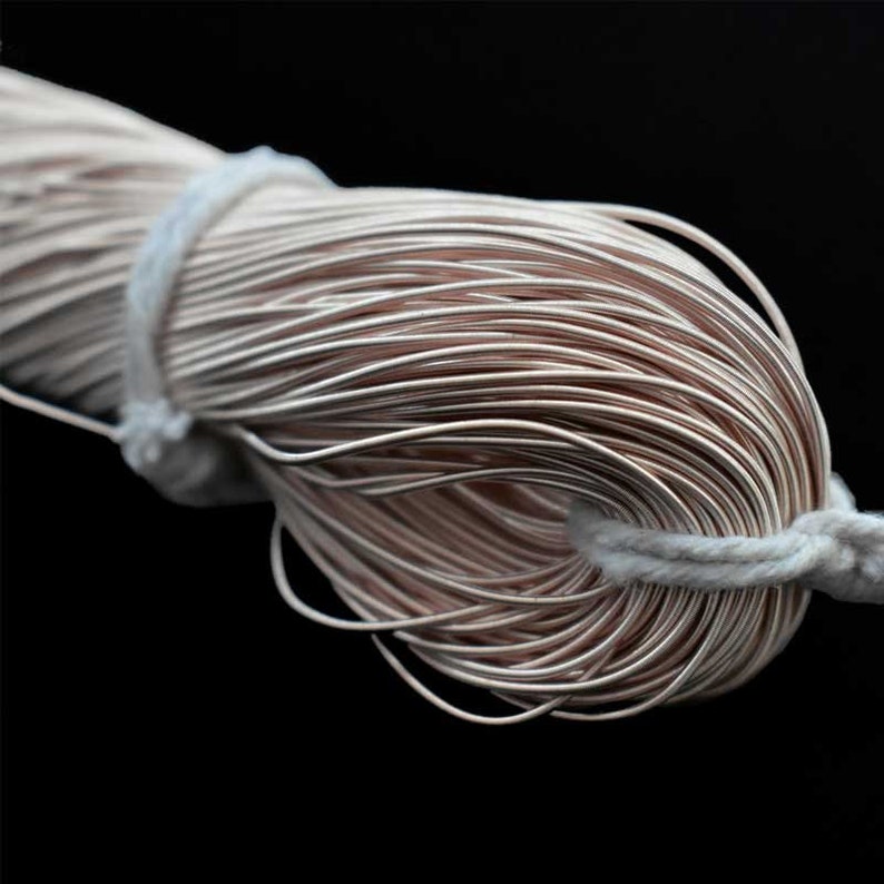 May include: Close-up of a bundle of thin, light brown wire, tightly wound and secured with a white cord. The wire has a metallic sheen, and the background is solid black.