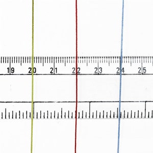 May include: Close-up of three colored threads: yellow-green, red, and light blue. A ruler is positioned behind the threads, with measurements in inches. The threads are vertical and parallel, showcasing their color and texture.