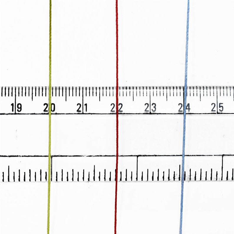 May include: Close-up of three colored threads: yellow-green, red, and light blue. A ruler is positioned behind the threads, with measurements in inches. The threads are vertical and parallel, showcasing their color and texture.