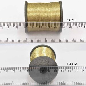 May include: Two spools of gold thread, one measuring 5 cm and the other 4.4 cm in diameter. The spools are on a white surface with a ruler.