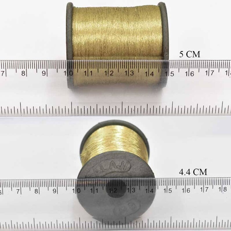 May include: Two spools of gold thread, one measuring 5 cm and the other 4.4 cm in diameter. The spools are on a white surface with a ruler.