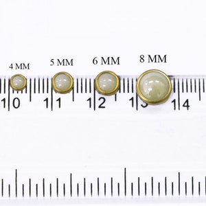 Op de afbeelding: Vier witte parelmoerachtige knopen met gouden randen, met een diameter van 4 mm, 5 mm, 6 mm en 8 mm, zijn op een liniaal uitgelijnd.