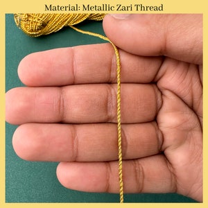 K&ouml;nnte beinhalten: Nahaufnahme eines goldenen, metallischen Zari-Garn, einer Art Stickgarn, das in einer Hand gehalten wird. Das Garn ist gedreht und hat eine gl&auml;nzende, metallische Oberfl&auml;che.