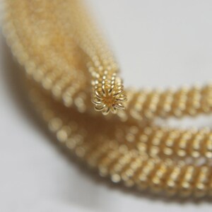 May include: A close-up of a gold chain made of small, linked beads. The chain is coiled and the end is visible, showing the intricate detail of the beadwork.