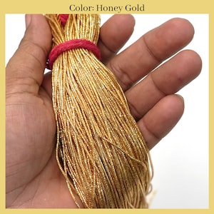 May include: A close-up of a bundle of gold metallic cord. The cord is thin and has a slightly textured surface. The cord is held in a hand, and the color of the cord is described as "Honey Gold".