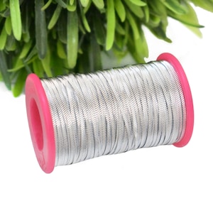 Pode incluir: Uma bobina de cordão metálico prateado com uma bobina de plástico rosa. O cordão está enrolado firmemente em torno da bobina.
