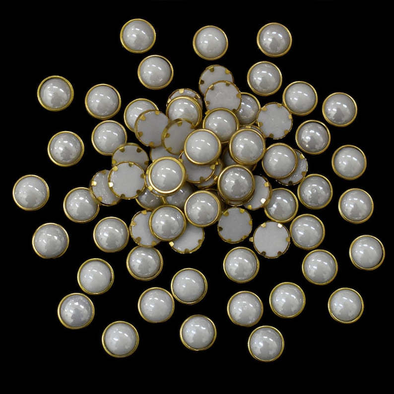 Op de afbeelding: Een grote stapel witte parelmoerachtige knopen met gouden metalen randen. De knopen zijn verspreid over een zwarte achtergrond.
