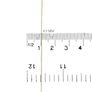 May include: A close-up of a gold cord or thread measured against a ruler. The ruler is marked in 1/2 inch increments and 0.5 mm increments. The cord is approximately 0.5 mm thick.