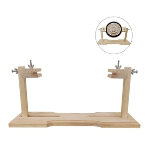 Puede incluir: Un soporte de madera para bastidor de bordado con base rectangular y dos soportes verticales. Cada soporte tiene una ranura horizontal y tuercas de mariposa ajustables para asegurar el bastidor. El soporte es de madera clara y está diseñado para sujetar un bastidor.