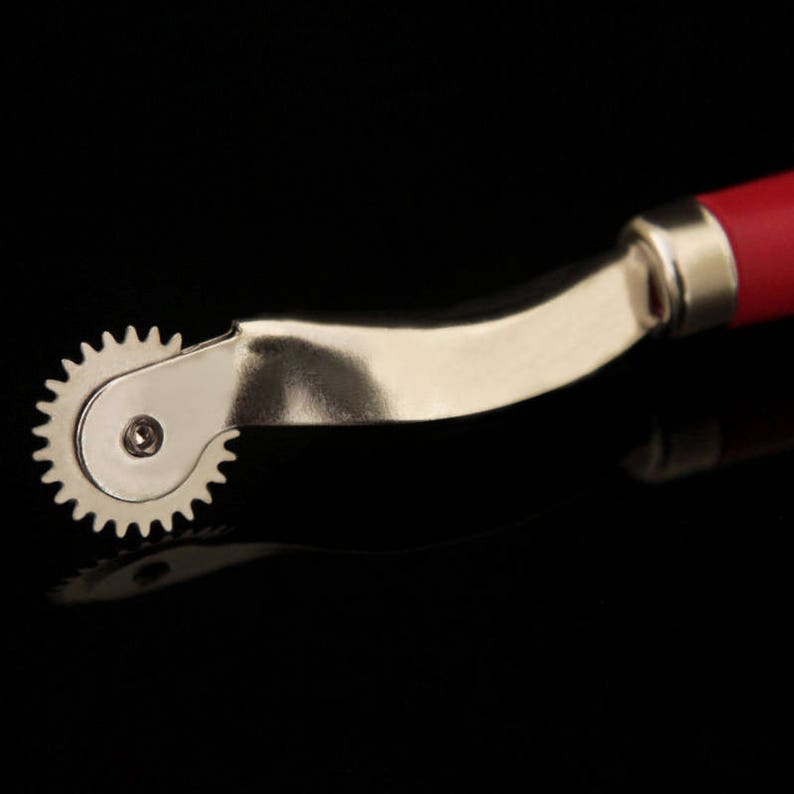 May include: A silver sewing tool with a red handle and a circular, toothed wheel. The tool is designed for creating even stitches in fabric.