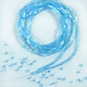 May include: A strand of light blue glass beads, with a square shape, arranged in a circle. The beads are scattered around the circle, creating a decorative effect.