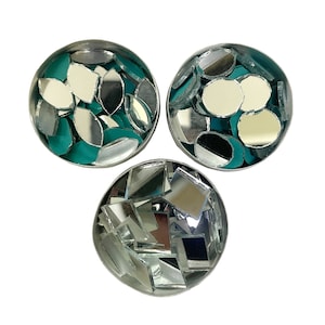 May include: Three small, round containers filled with reflective, silver-colored mirror pieces. The mirror pieces are various shapes, including leaf-like, round, and square. Each container has a teal-colored base. The background is plain white.