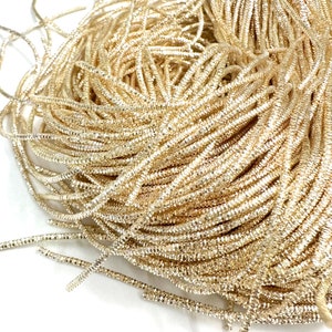 May include: A close-up of a spool of gold metallic cord. The cord is made of small, shiny squares that are woven together to create a shimmering effect.