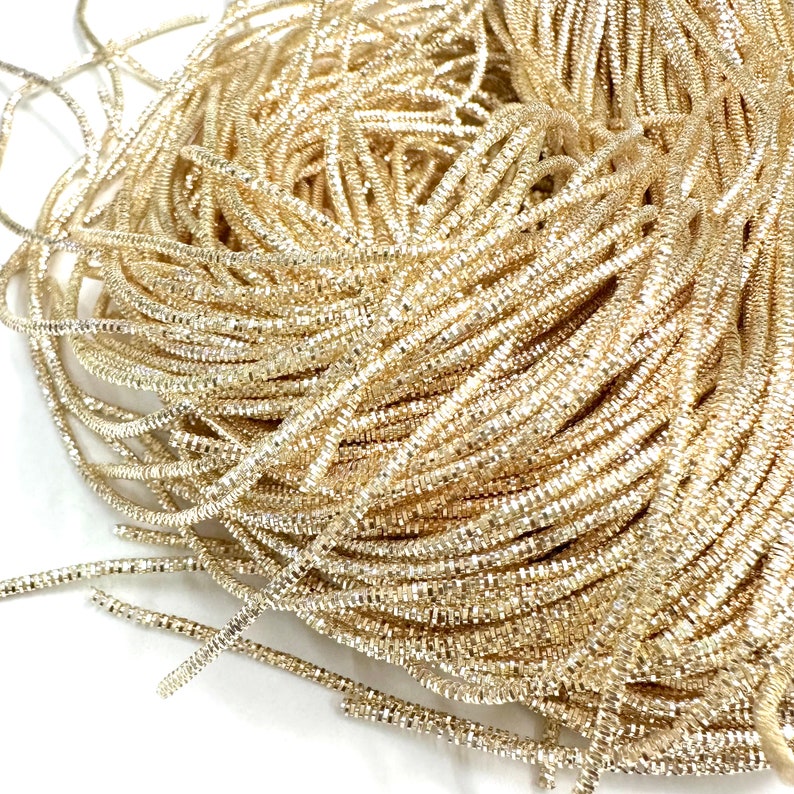 May include: A close-up of a spool of gold metallic cord. The cord is made of small, shiny squares that are woven together to create a shimmering effect.