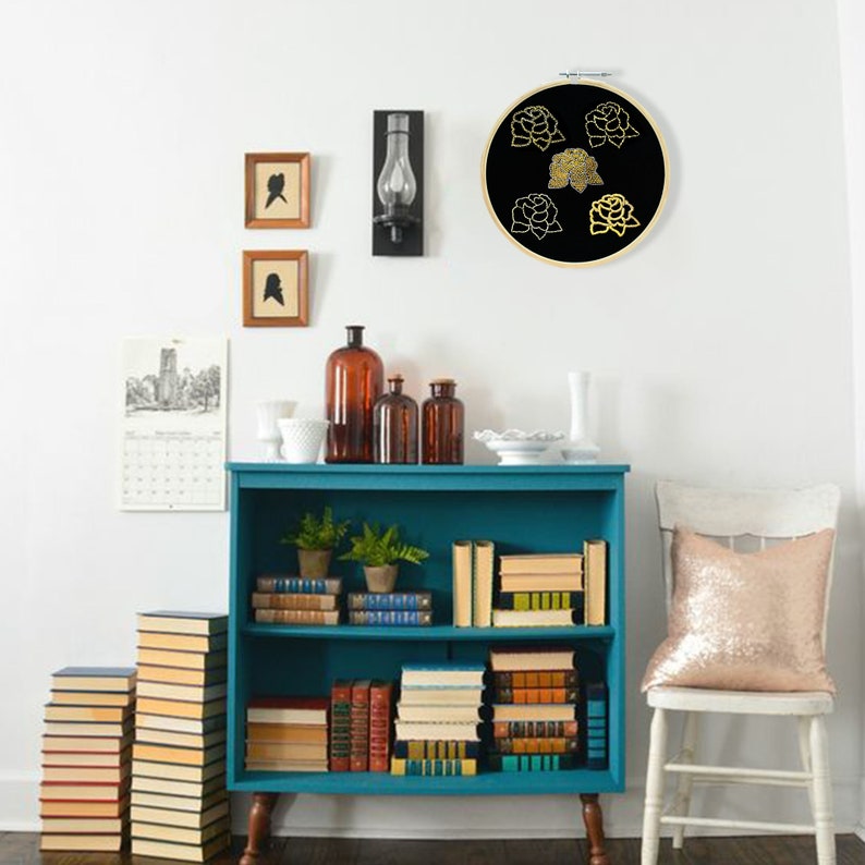 May include: A black and gold embroidery hoop with a floral design. The hoop is hanging on a white wall above a teal bookshelf with books and decorative items.