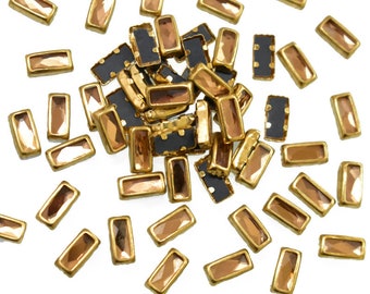 May include: A pile of gold-colored rectangular nail rhinestones with a clear, faceted center. The rhinestones are scattered on a white background.