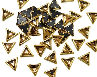 Puede incluir: Una dispersión de piedras de cristal triangulares con borde dorado y un centro transparente de color ámbar. Las piedras de cristal están dispersas sobre un fondo blanco.