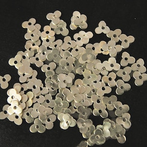 Puede incluir: Un montón grande de lentejuelas de metal de color plateado en forma de flor. Las lentejuelas están esparcidas sobre un fondo negro.